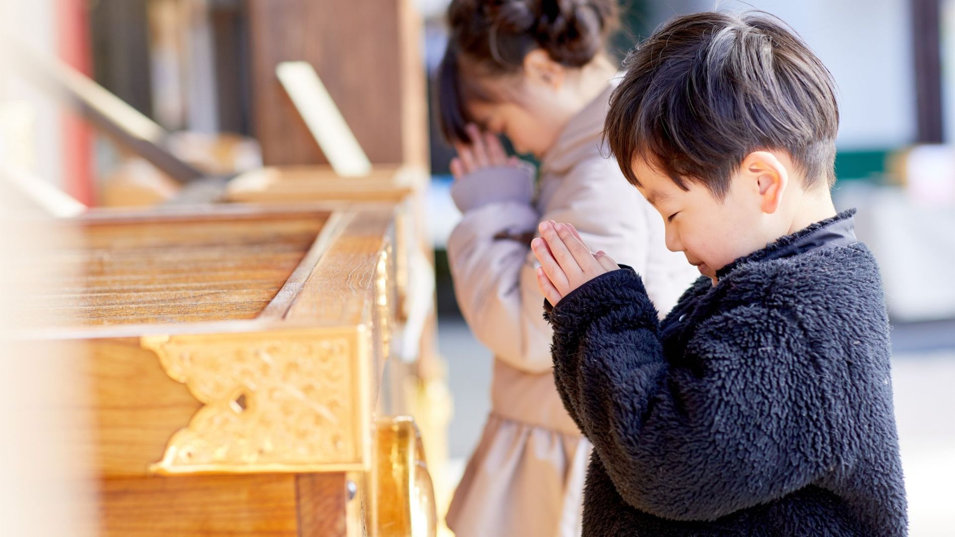 Two children with hands clasped in prayer near a wooden structure outdoors.