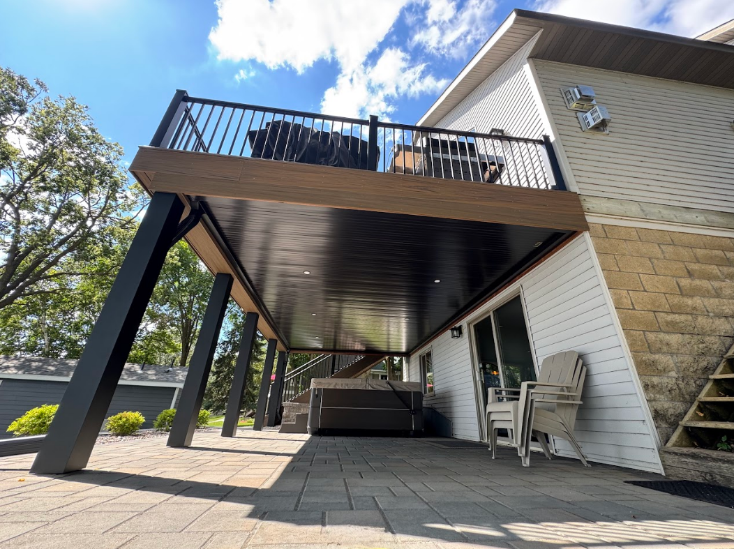 A house with a black-and-brown deck, supported by black beams, overlooks a stone patio.