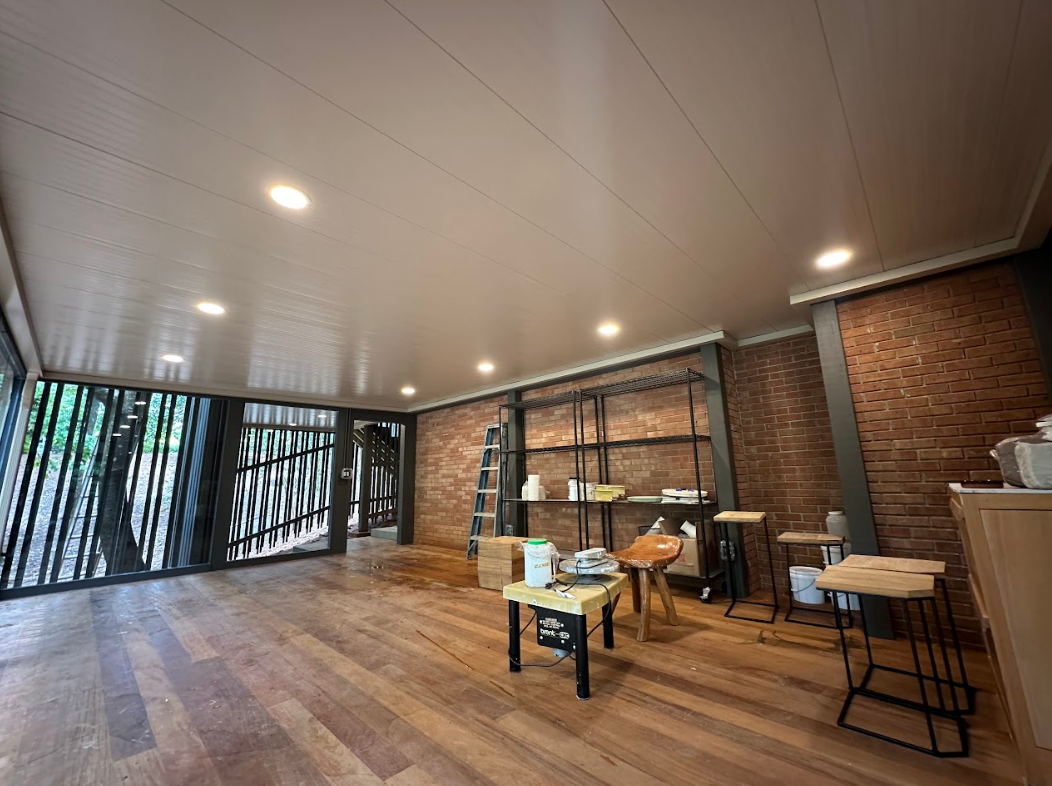 Spacious room with wood floor and brick walls, featuring shelves, tables, and a view of trees.