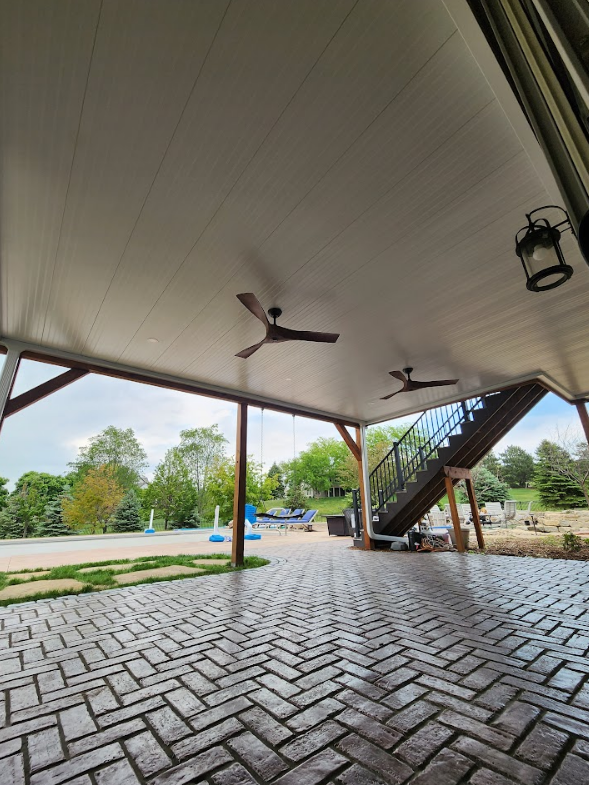 Patio with brick-patterned floor, wooden supports, ceiling fans, and staircase leading up. Overlooking a pool.