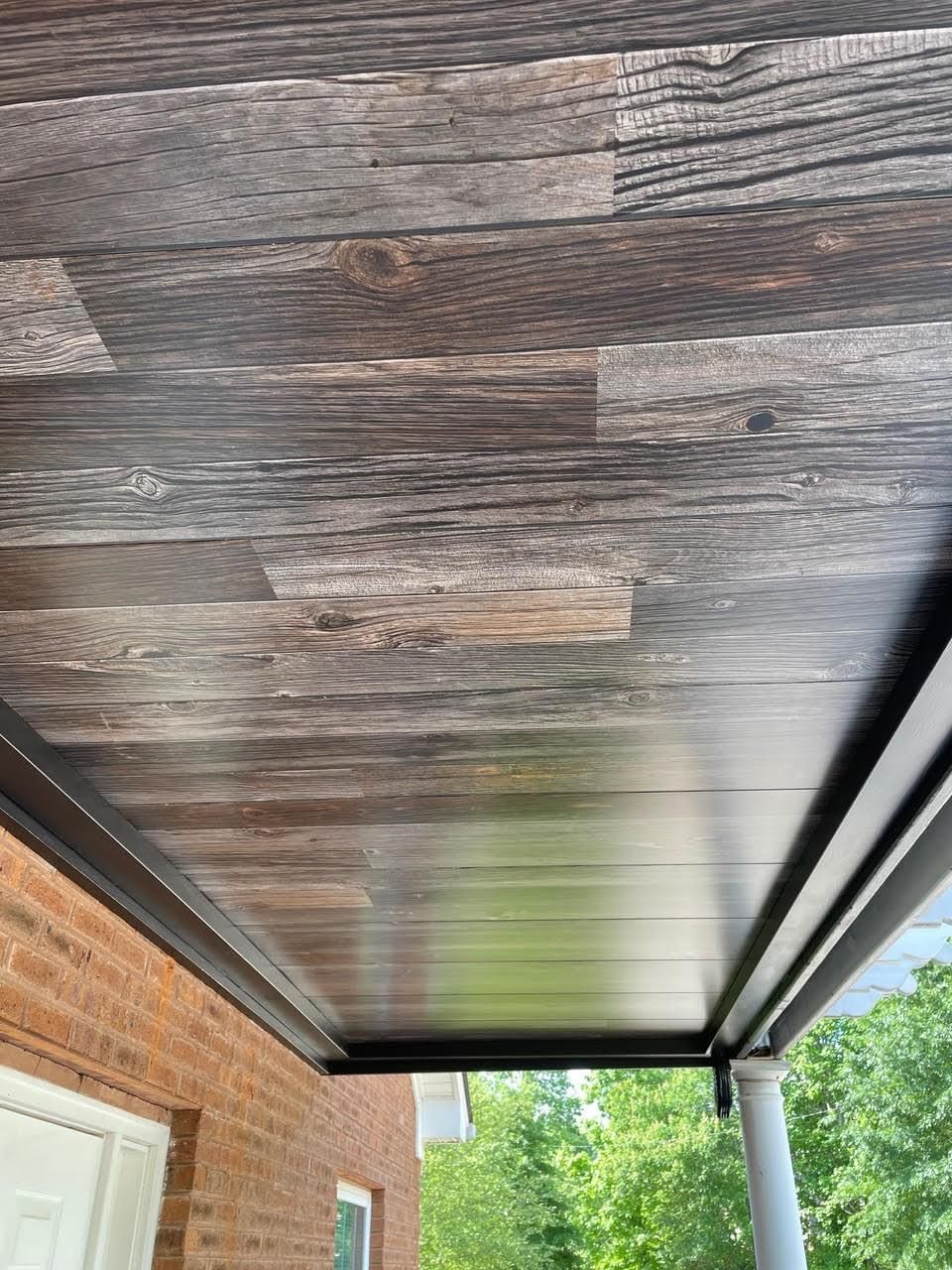 Wood-look ceiling on a porch with dark trim, viewed from below. The brick house and trees are visible.
