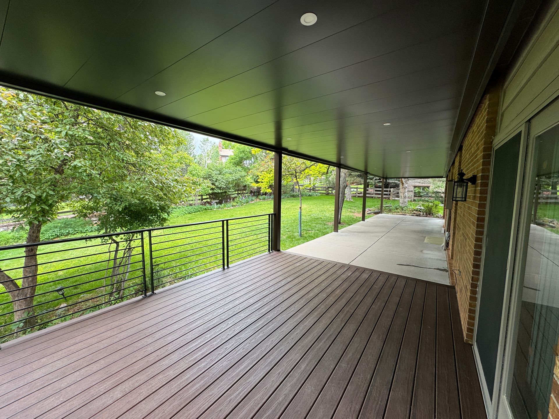 Covered wooden deck overlooking a grassy yard with trees, black railing, and a dark ceiling.