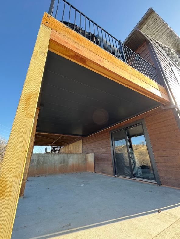 View from below of a wooden deck with black underside, overlooking a concrete patio area and building exterior.