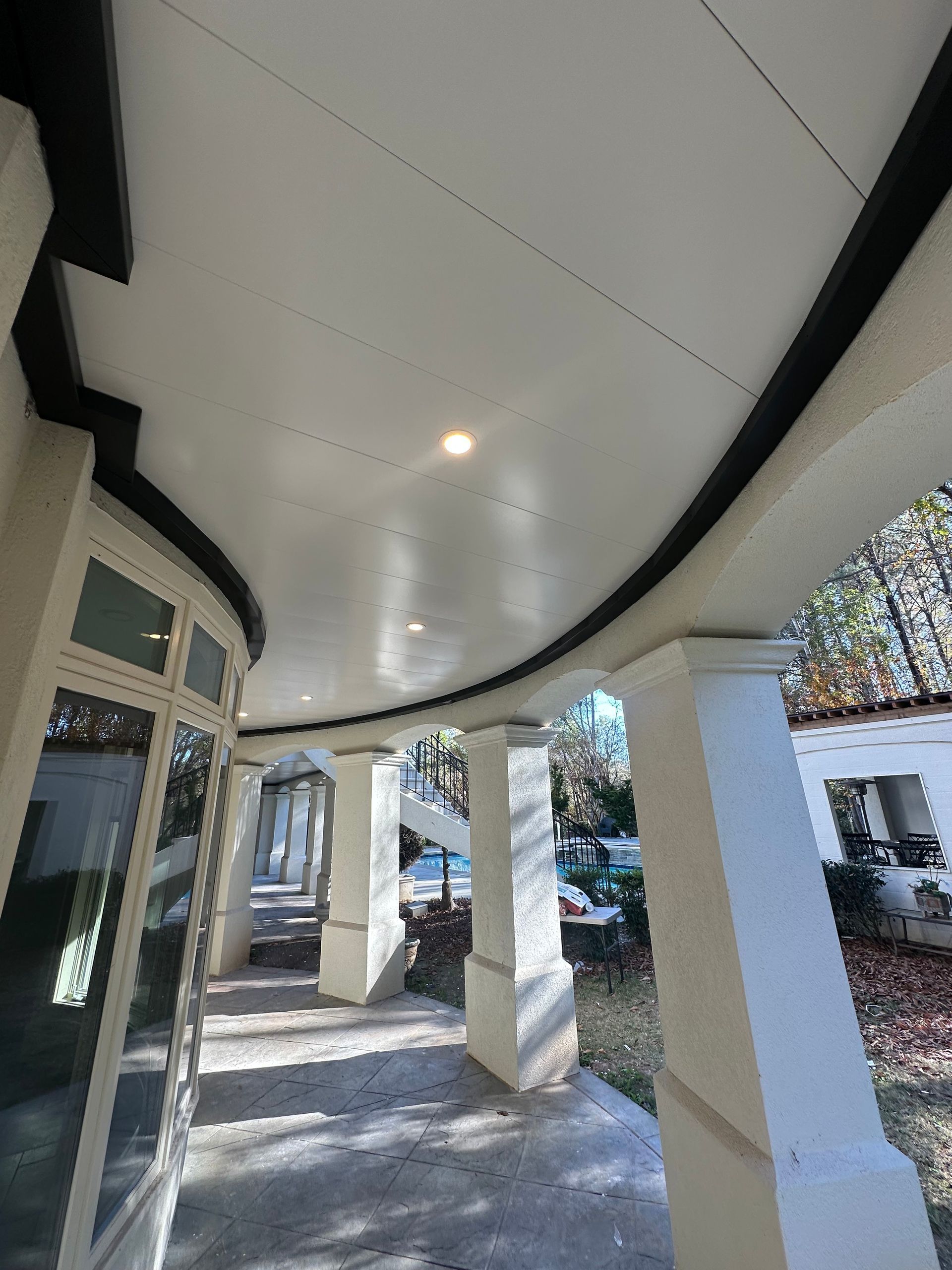 Curved white porch roof with black trim, pillars, and a distant view of a yard and building.