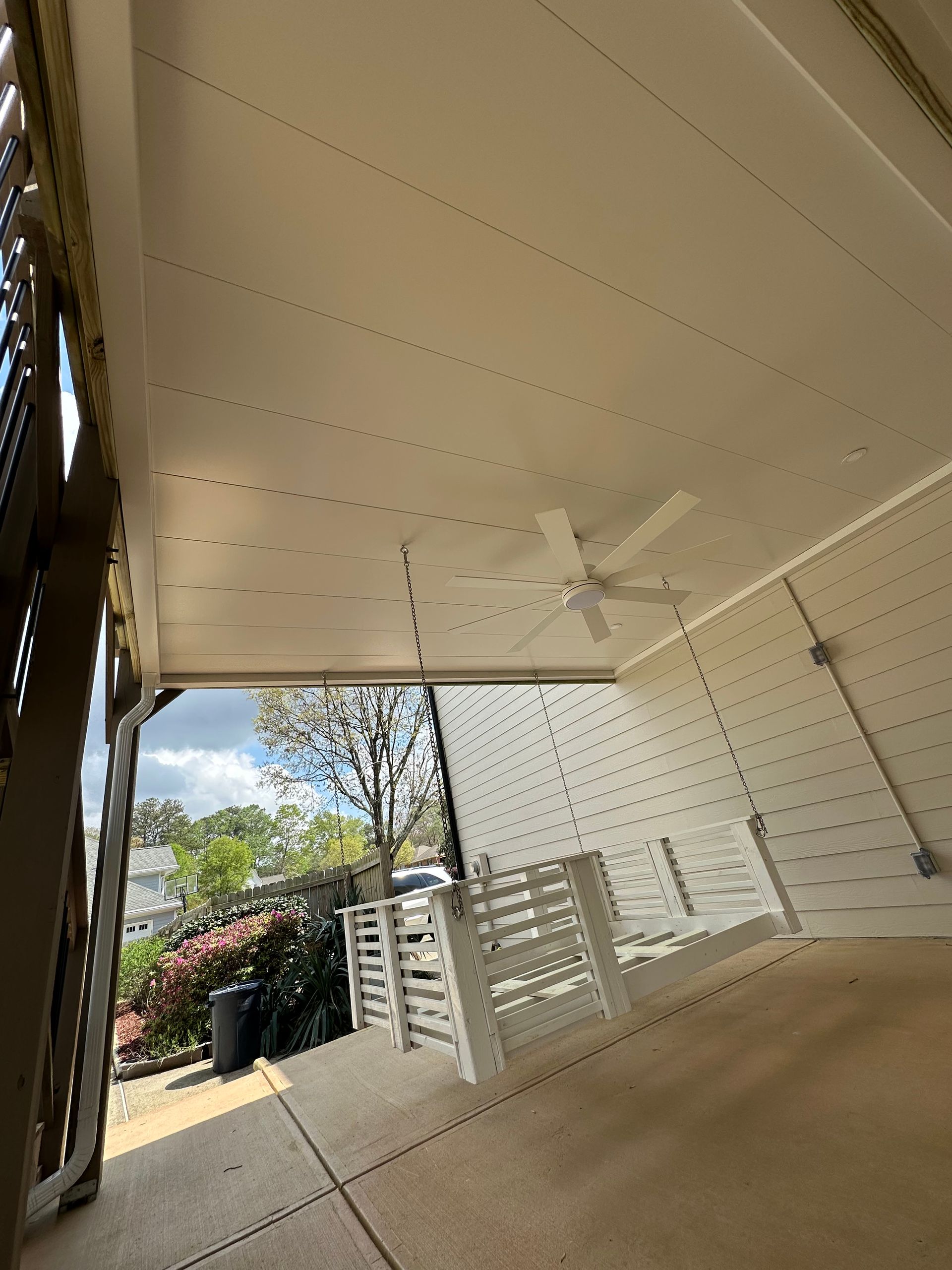 White porch with ceiling fan, white paneled wall, and a view of trees and a street outside.