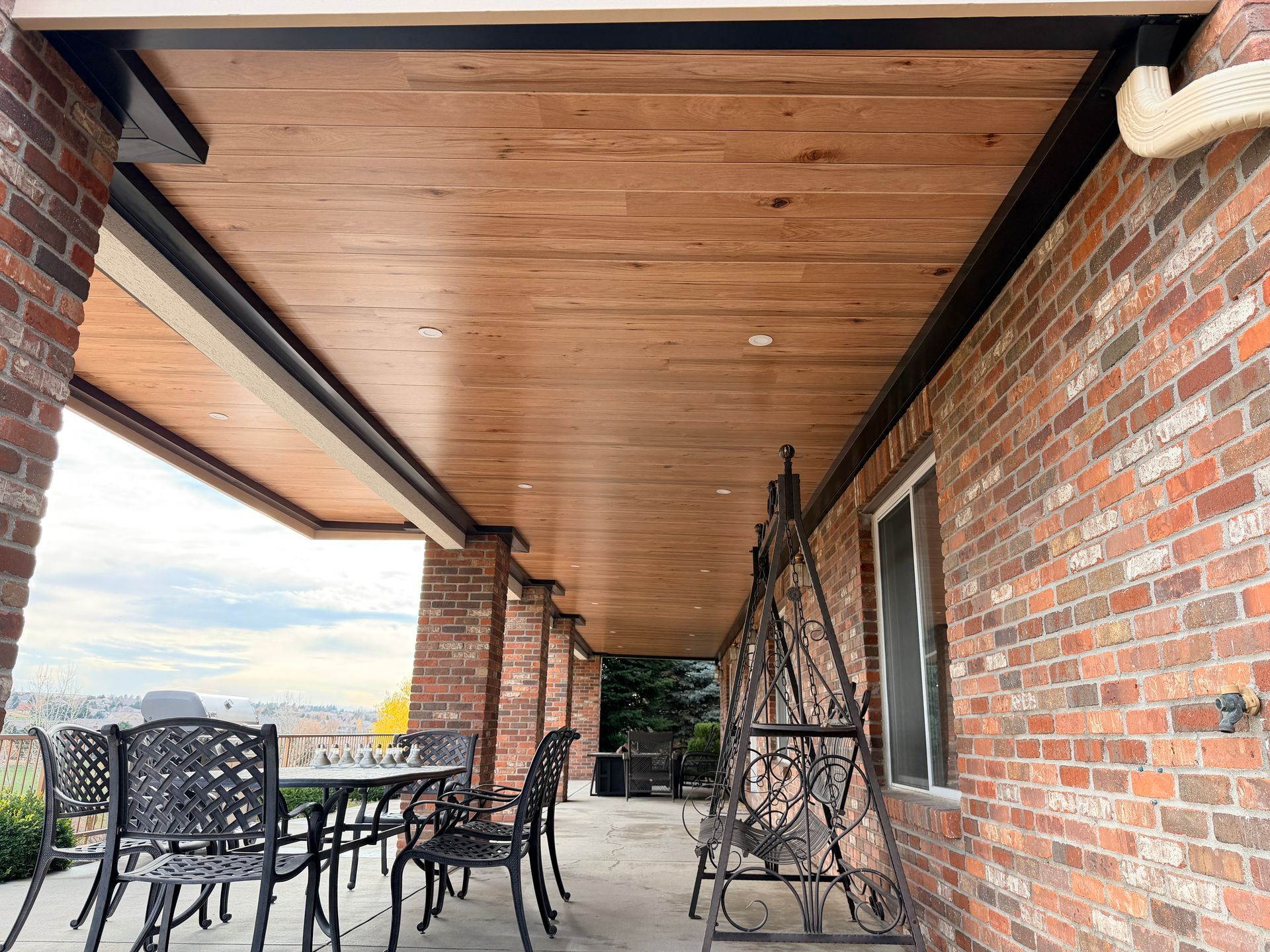 A covered brick porch with a wooden ceiling, a metal table and chairs, and a decorative wrought iron bench.