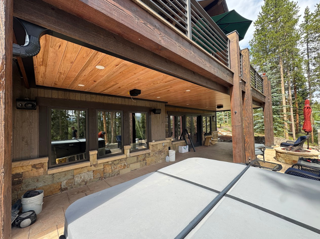 Outdoor deck with hot tub, windows, and wood ceiling, surrounded by trees.