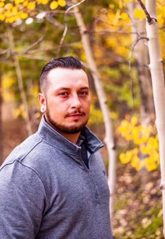 Man with a beard and gray sweater in a forest, looking toward the camera. Yellow and white trees are in the background.