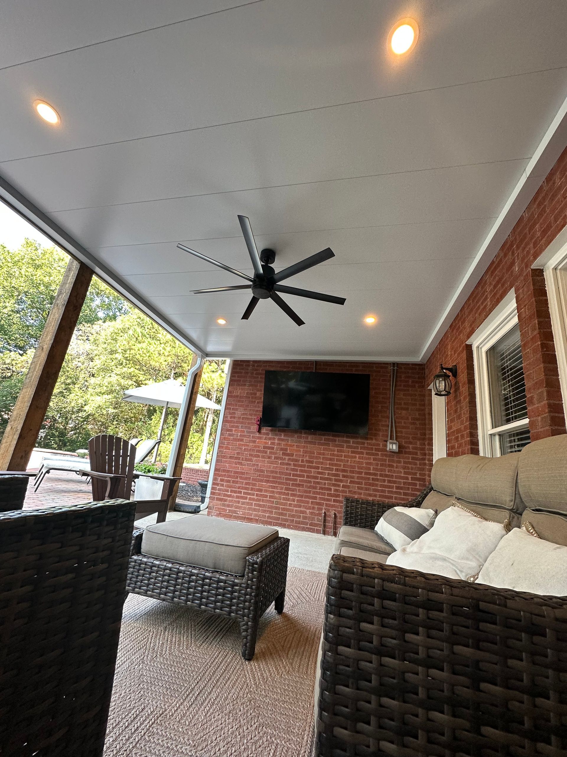 Covered outdoor patio with a TV, ceiling fan, and brick wall. Wicker furniture on a patio with recessed lighting.