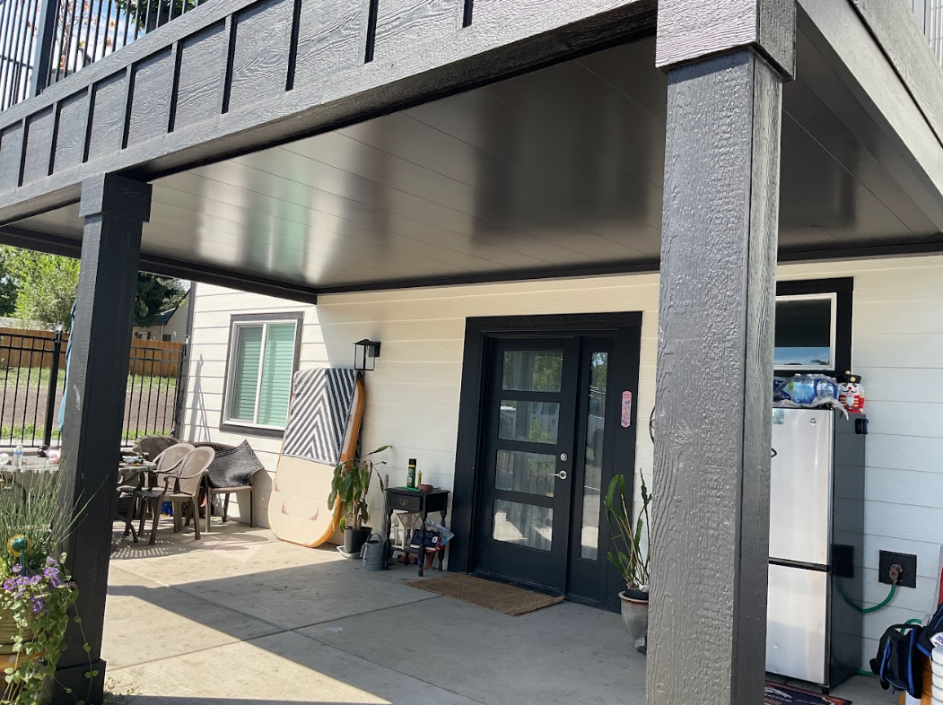 Black-framed patio cover over a white building with black doors. Outdoor seating and plants are visible.