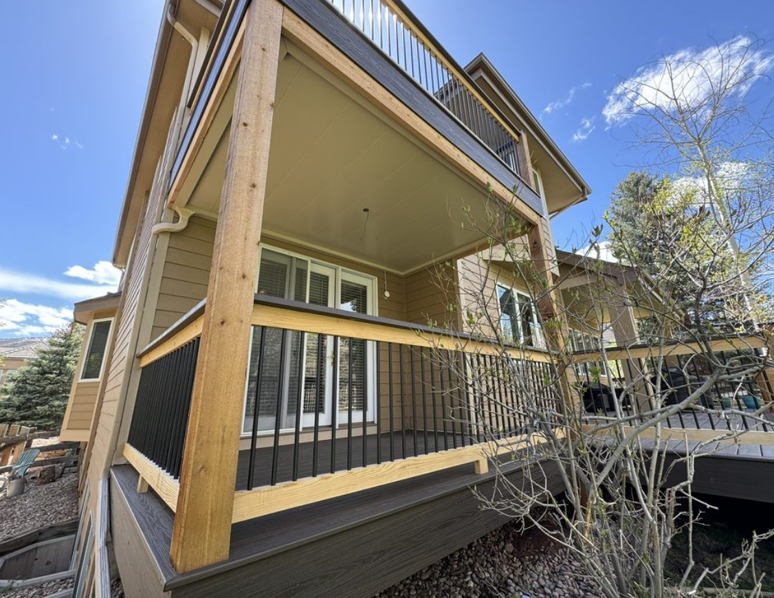 Two-story house with deck, brown and tan exterior, black railing. Sunny day.