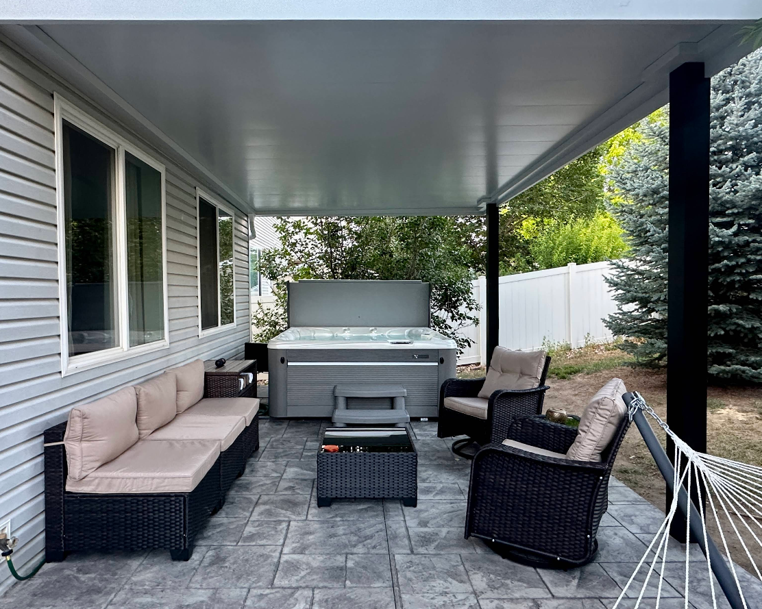 Patio with seating, hot tub under a covered roof. Gray siding and concrete with black accents.