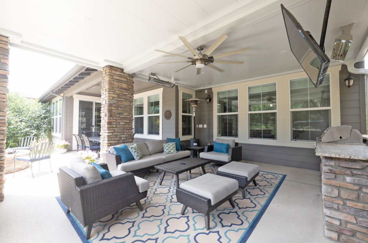 Covered patio with outdoor furniture, including sofa, chairs, ottomans, and rug. Features TV, ceiling fan, and stone accents.