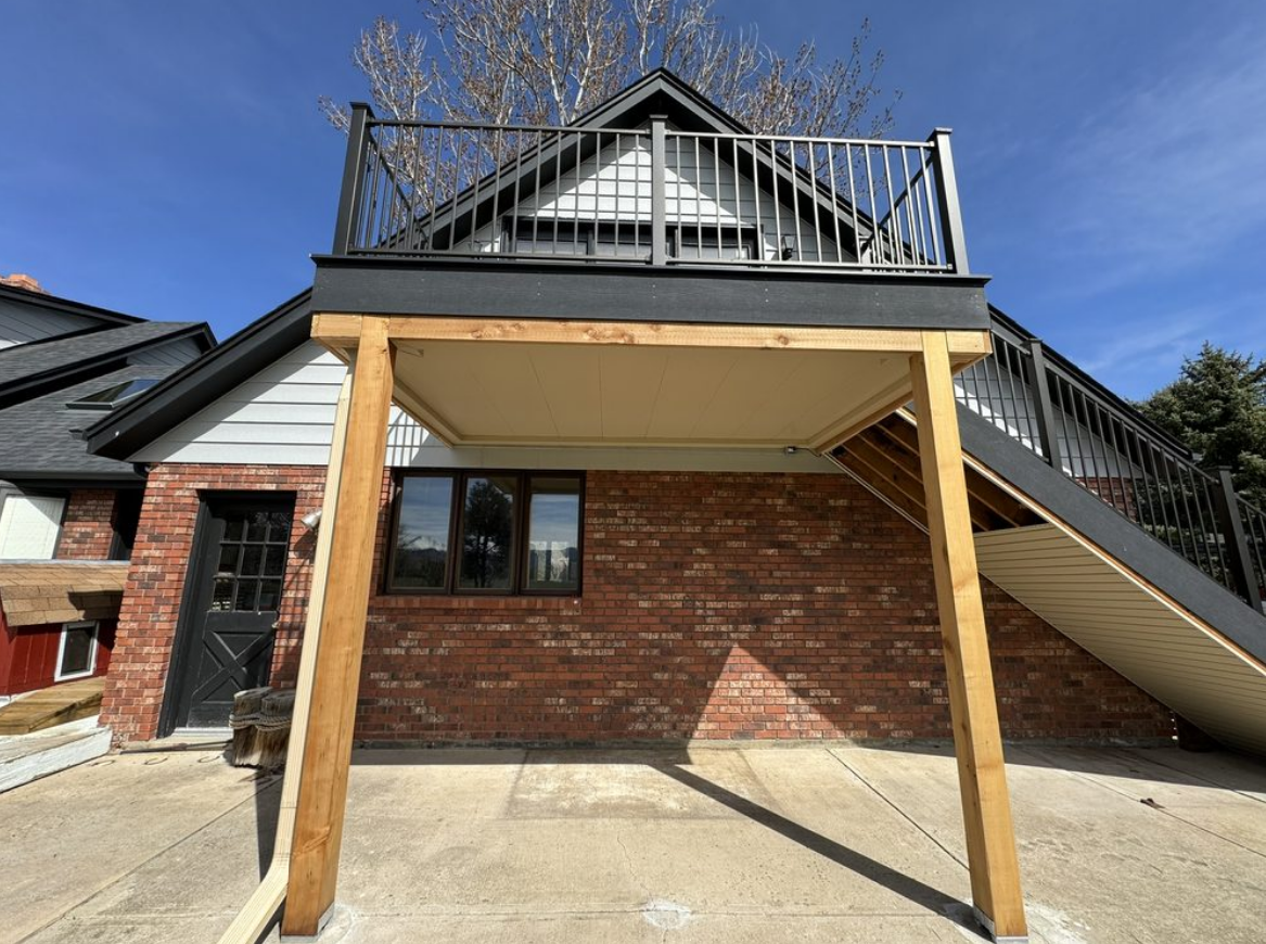 Exterior view of a building with a deck on top. A porch is beneath the deck, supported by wooden beams.