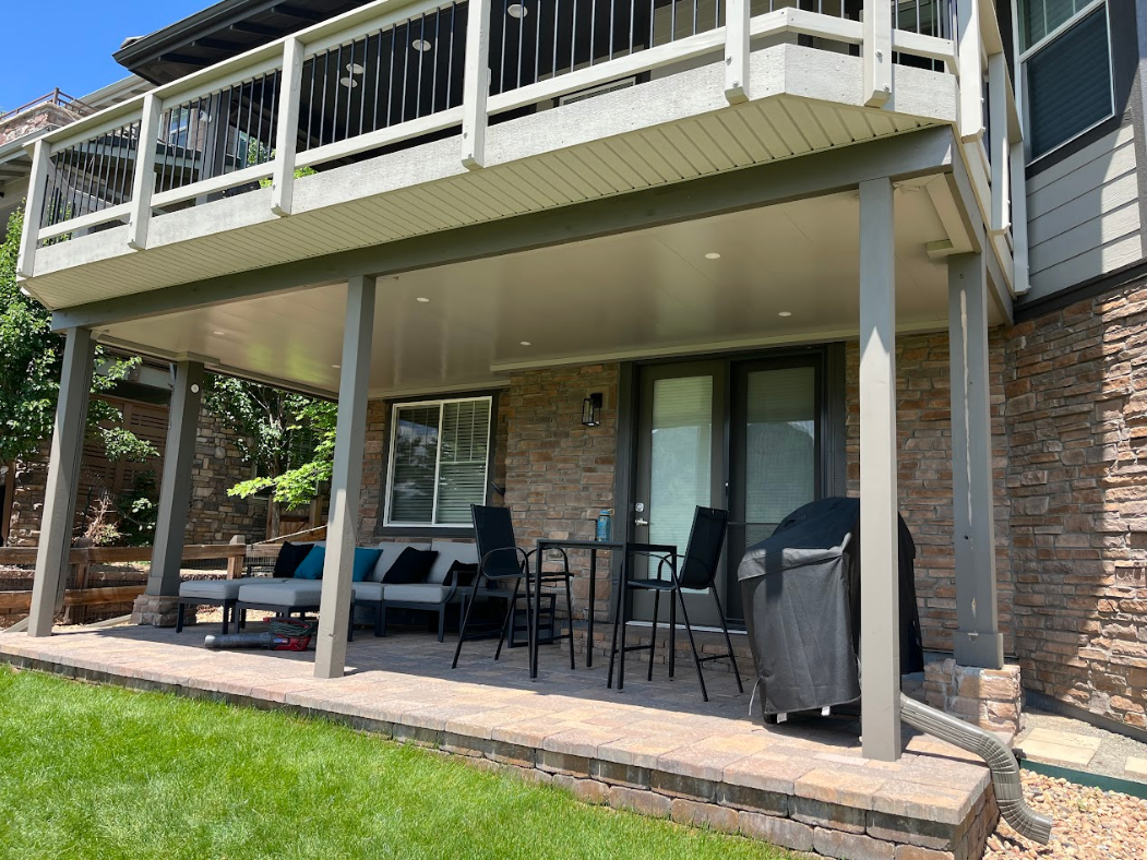 Covered patio under a deck with outdoor furniture, adjacent to a brick house and green lawn.