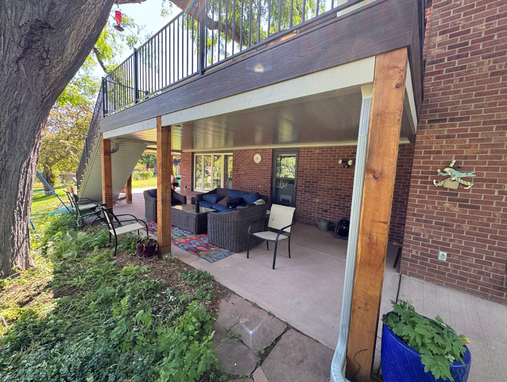 A covered outdoor patio with seating under a second-story deck, brick wall.