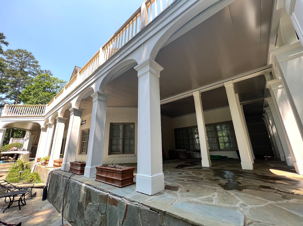 White colonnaded patio with stone flooring and a railing, with greenery in the background.
