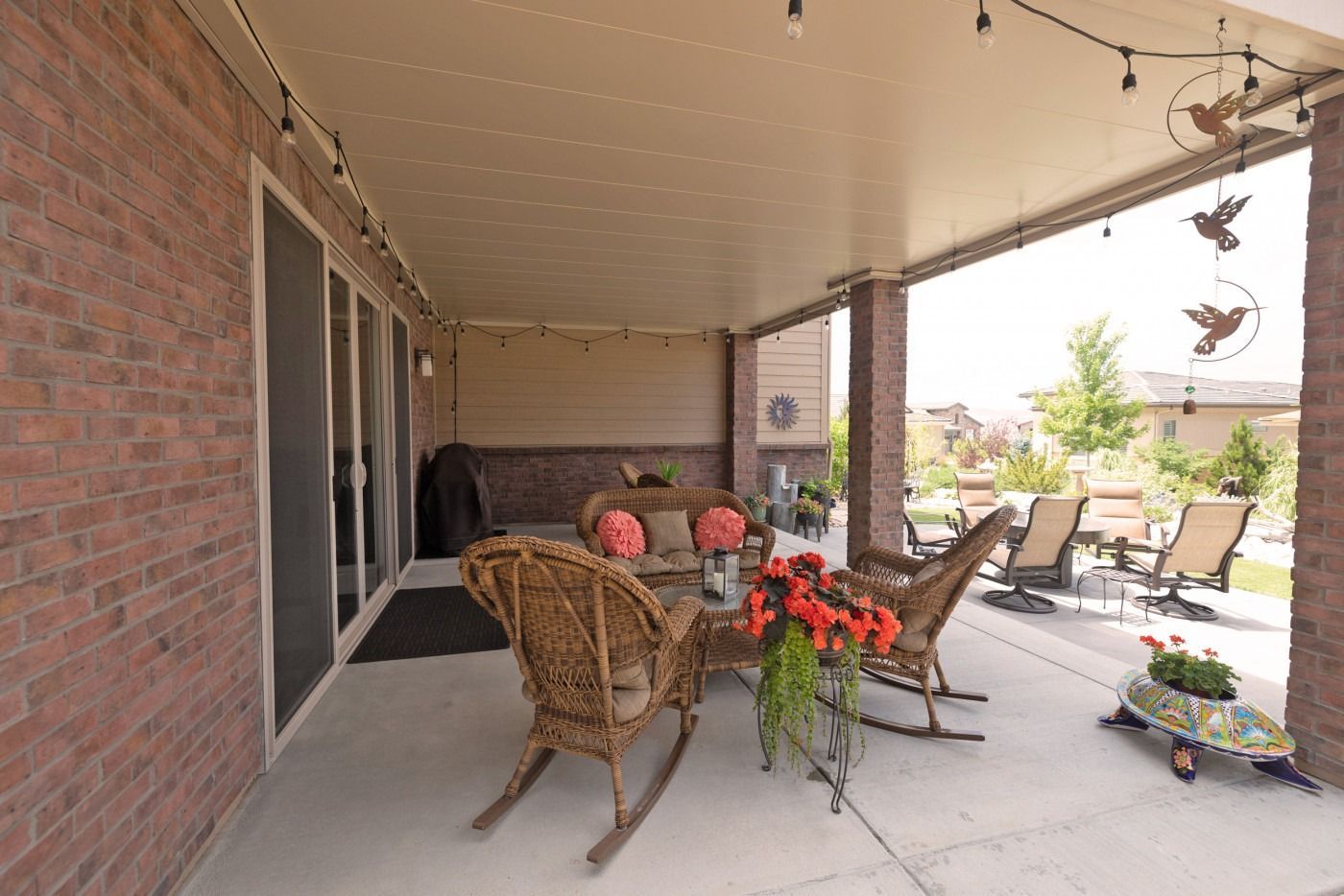 Covered patio with wicker furniture, red brick, and string lights overlooking a sunny yard.