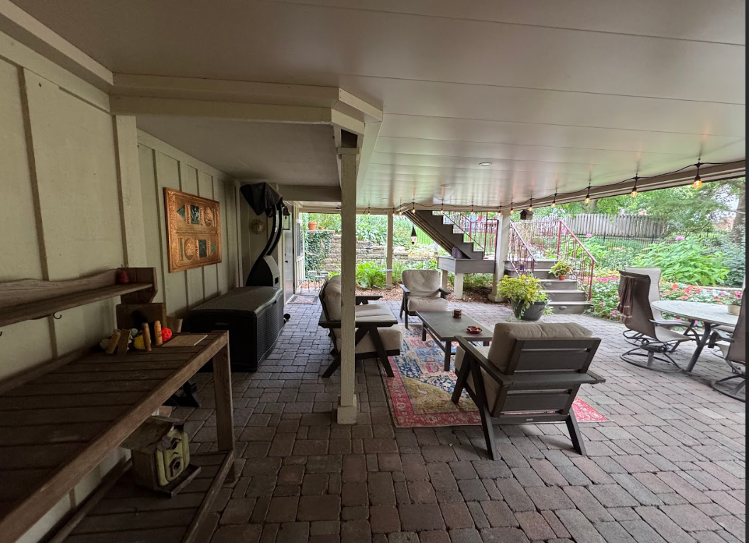 Covered brick patio with outdoor furniture, wood burner, and stairs leading to garden.