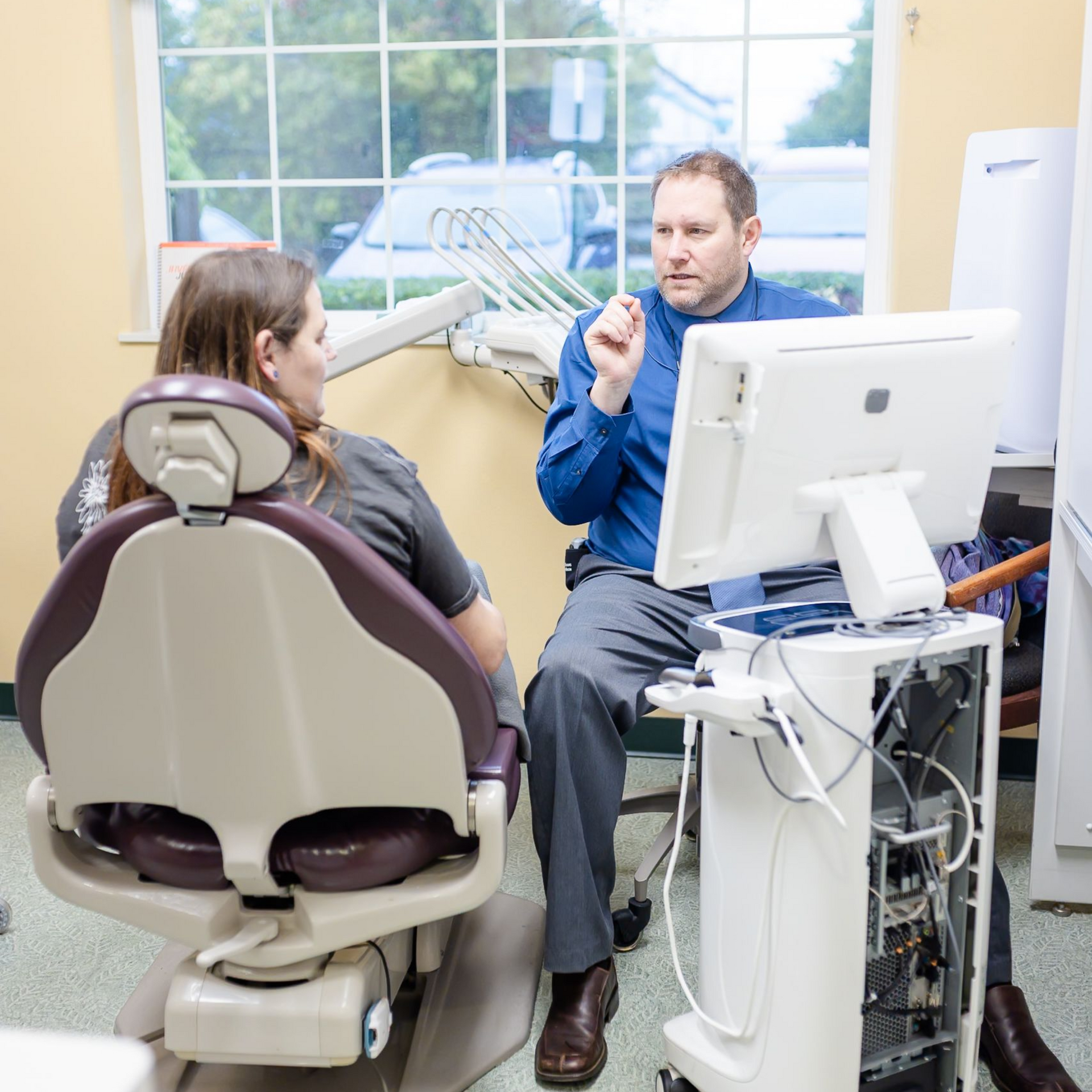 Dentist in blue shirt pointing at screen, talking to patient in chair. Dental office setting, window in background.