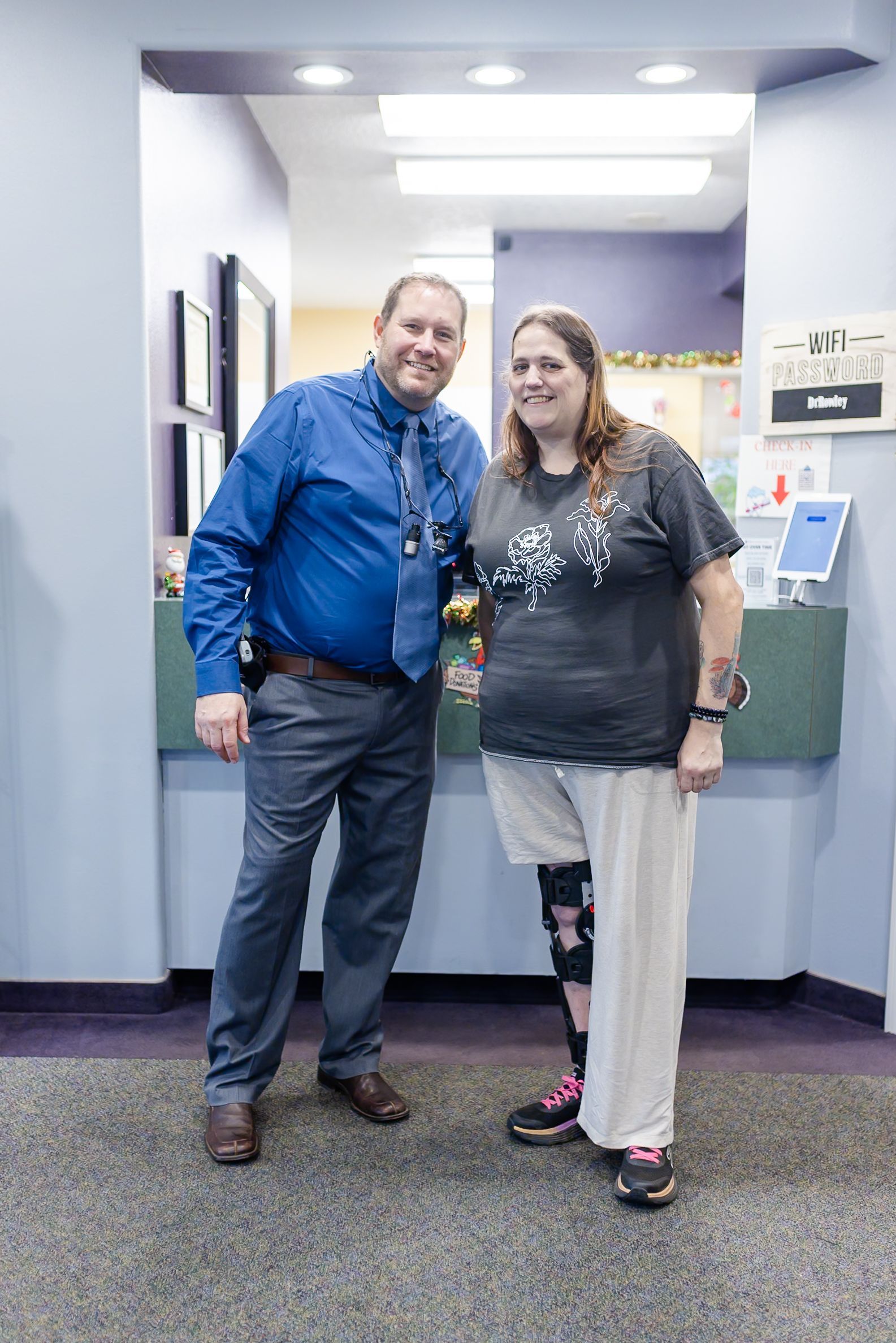 Man in blue shirt and woman with leg brace smile by a reception desk.