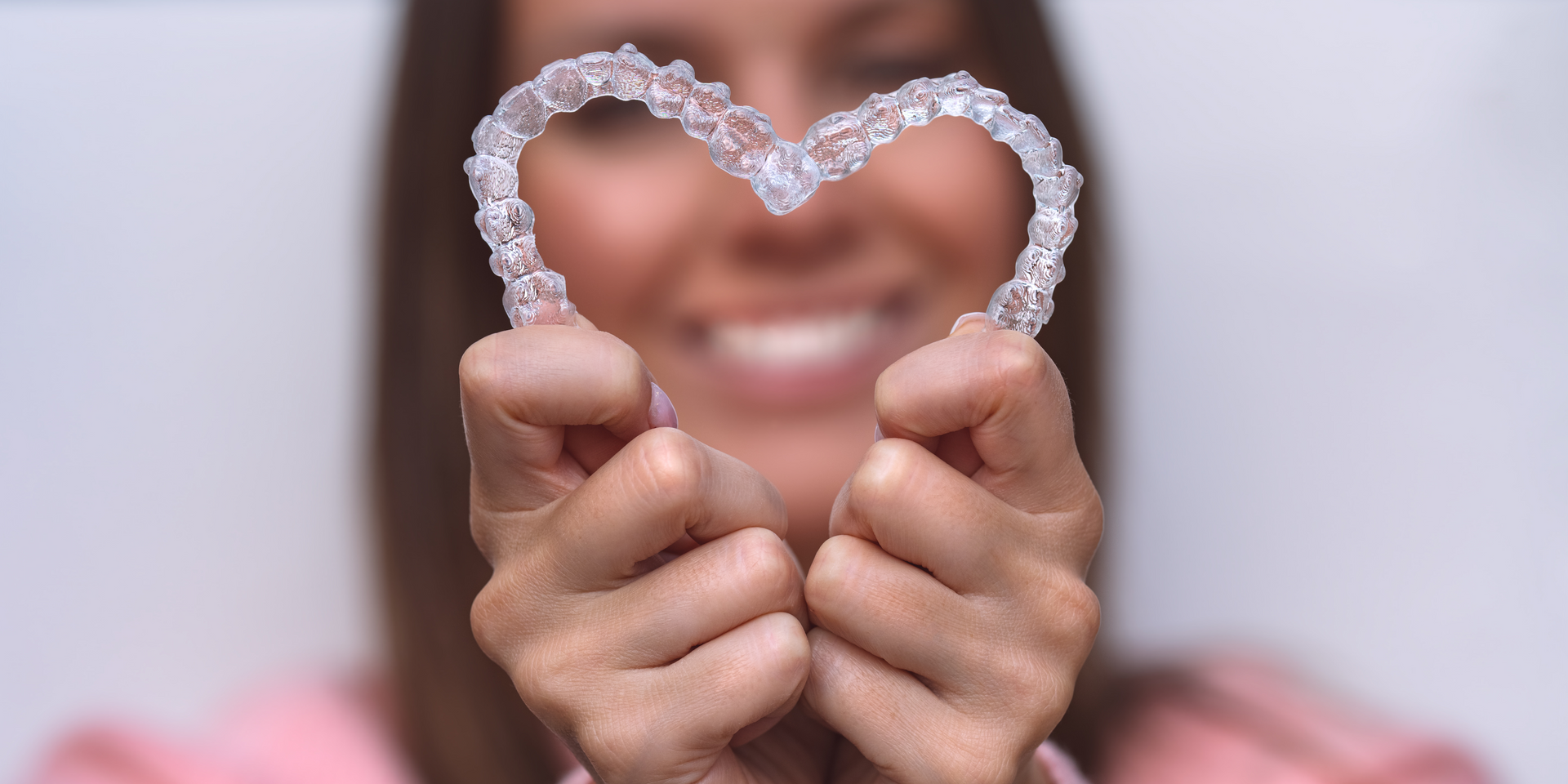 Woman holding clear dental aligners in the shape of a heart, smiling, with blurred background.