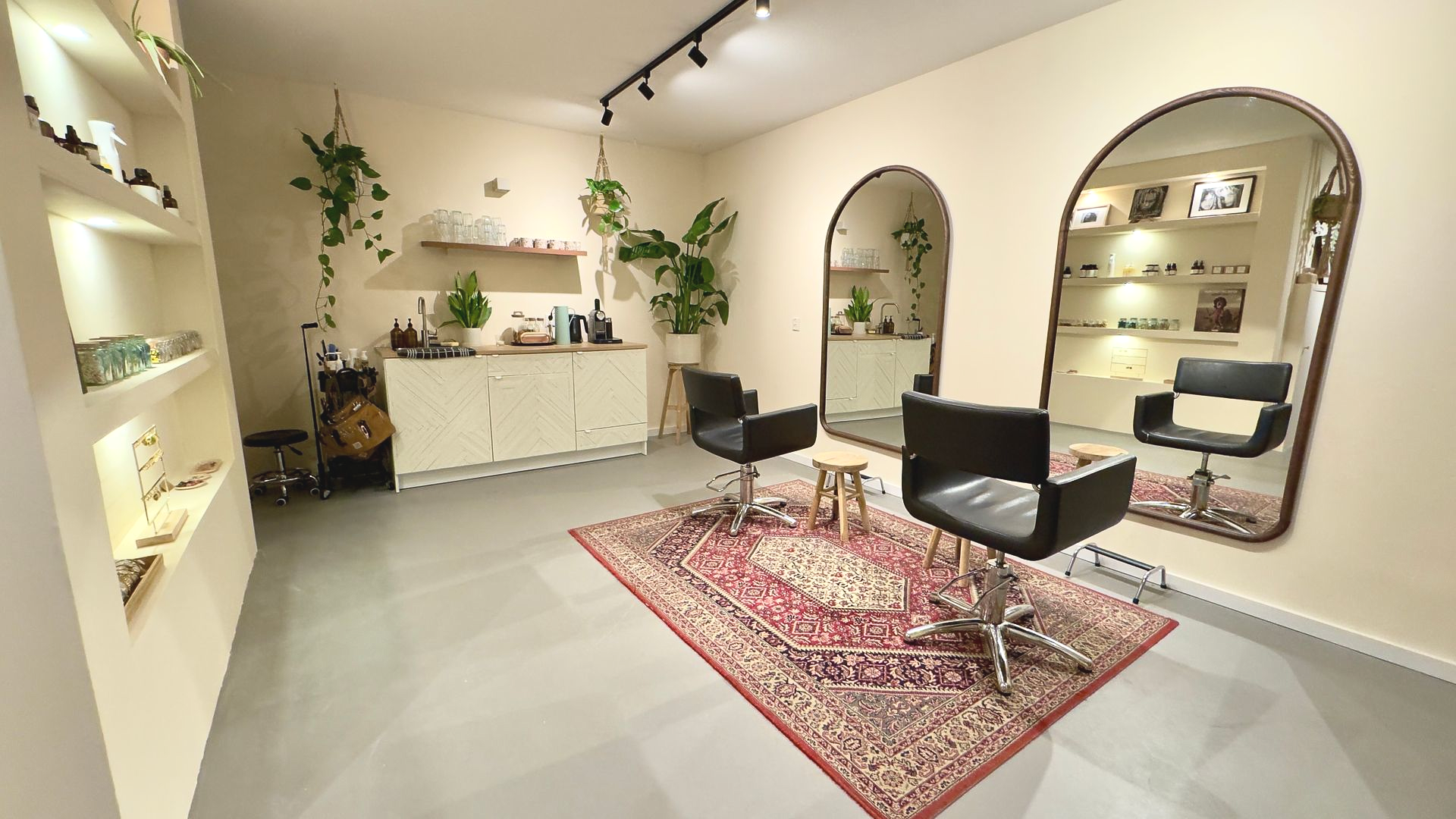 Interieur van een kapsalon met twee grote spiegels, stoelen en een Perzisch tapijt. Planten en planken met producten zijn zichtbaar.
