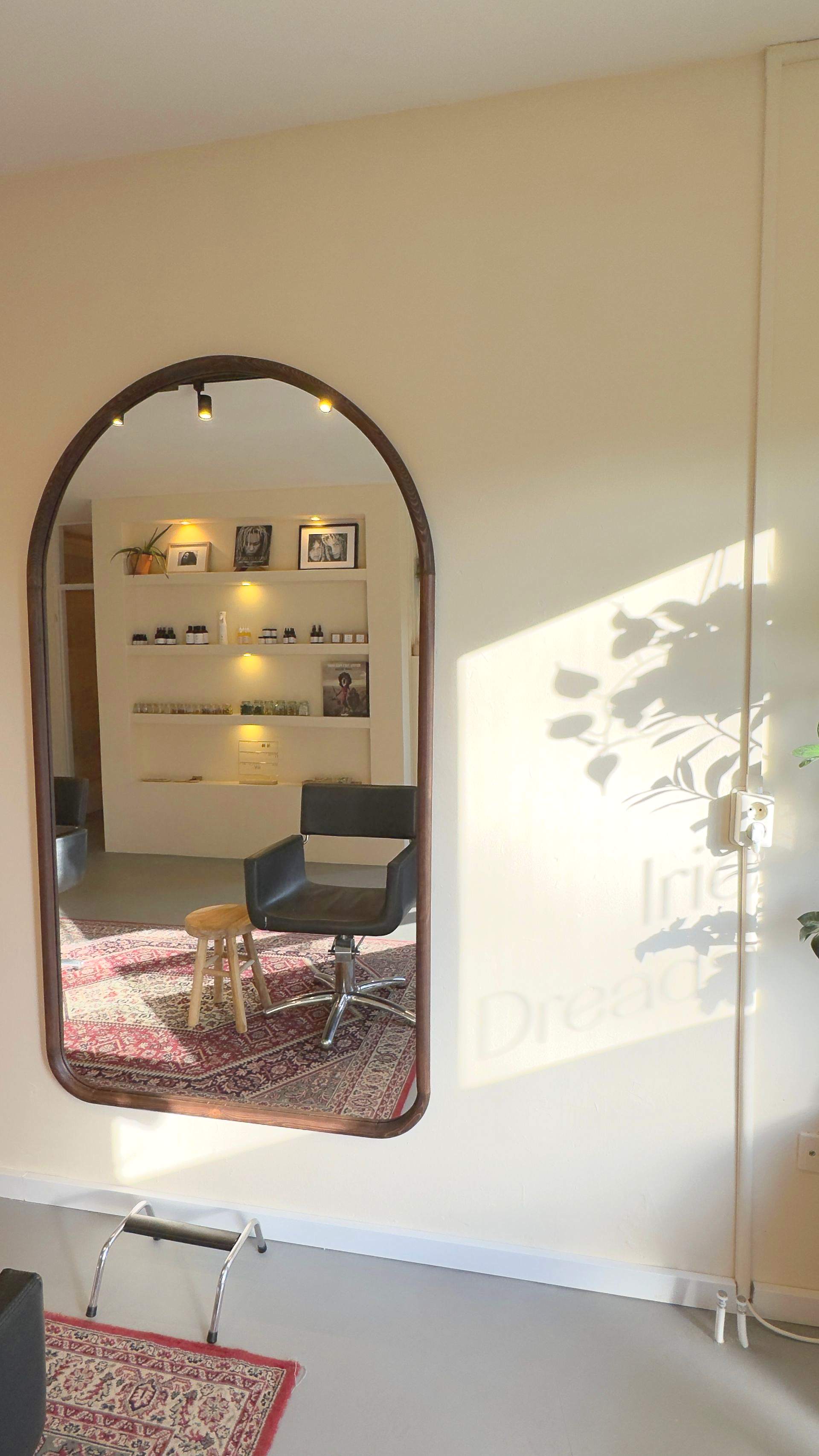 Een grote, gebogen spiegel weerspiegelt een interieur: stoelen, een vloerkleed en planken met tentoongestelde voorwerpen. Zonlicht verlicht de muur.