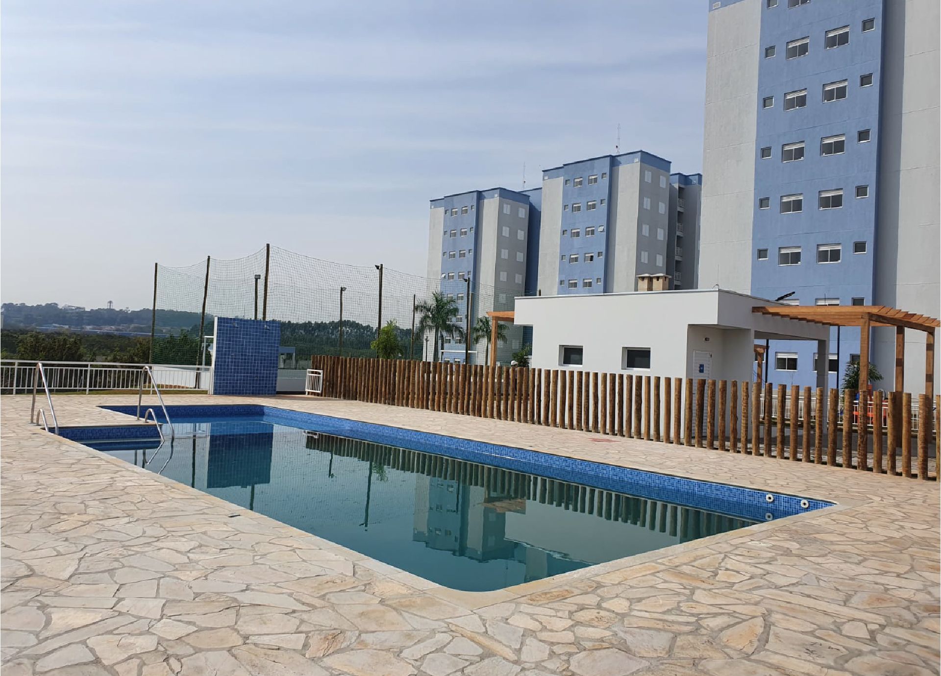 Piscina com água azul, cercada por pedras e uma cerca de madeira, ao lado de prédios de apartamentos sob um céu nublado.