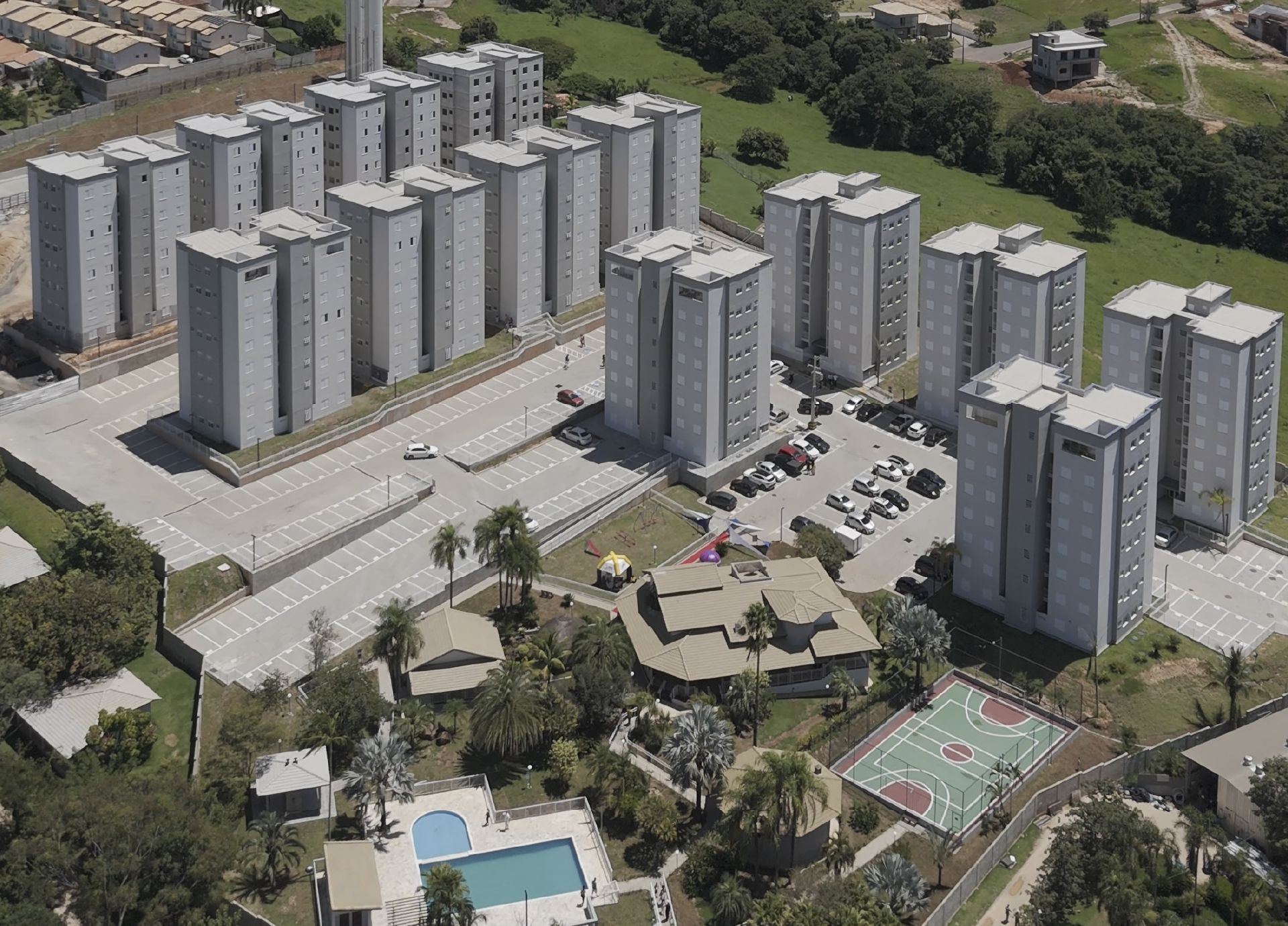 Vista aérea de muitos prédios altos e cinzas de apartamentos em um complexo, com piscina, quadra esportiva e área verde.
