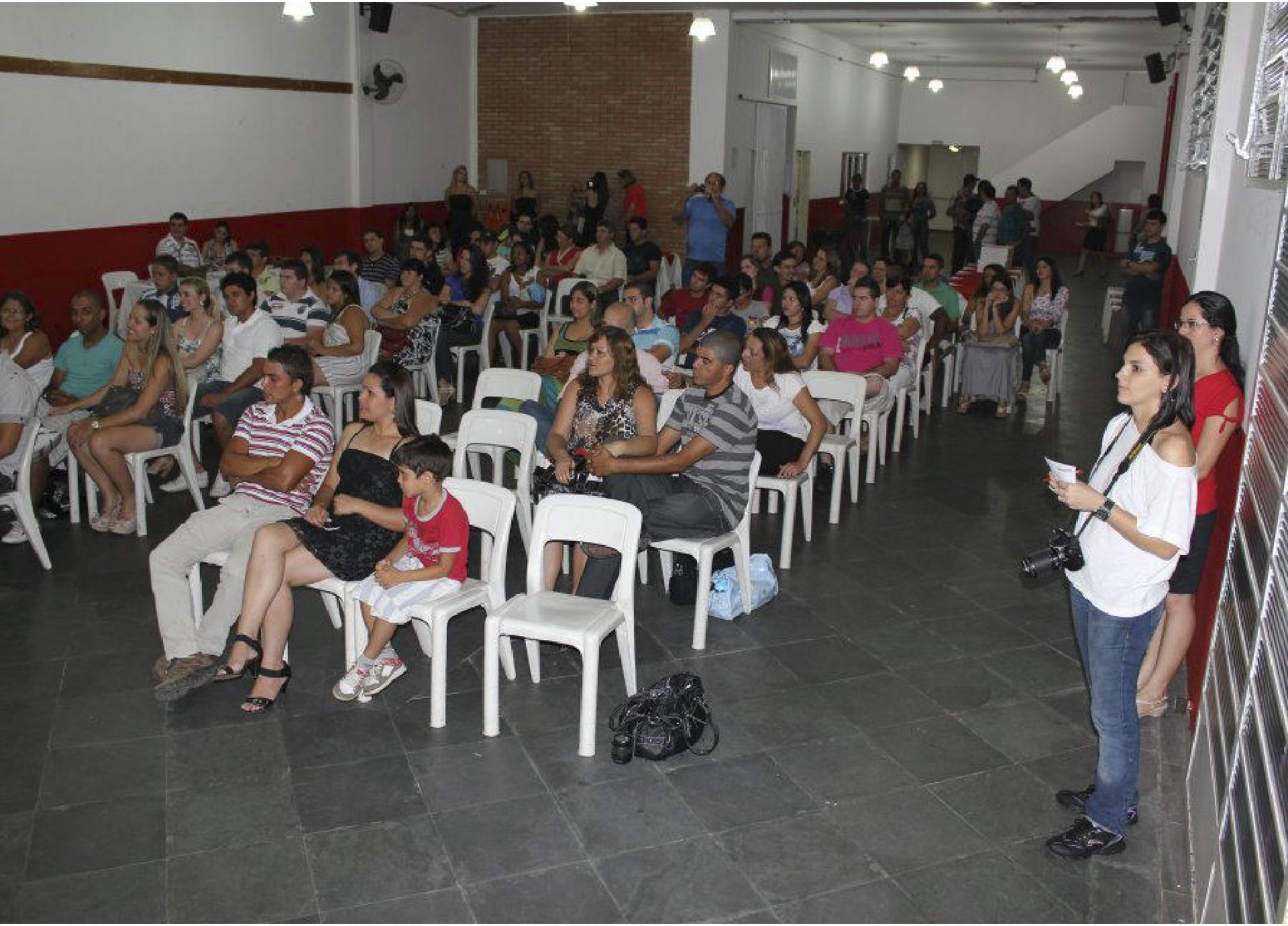 Público sentado em fileiras em um evento. Uma mulher com uma câmera em pé ao lado, tirando fotos.
