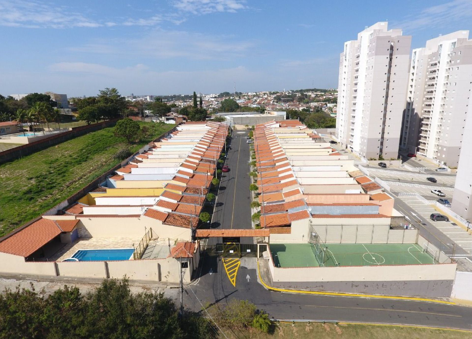 Vista aérea de um condomínio fechado com sobrados, piscina, quadra de tênis e prédios altos de apartamentos.