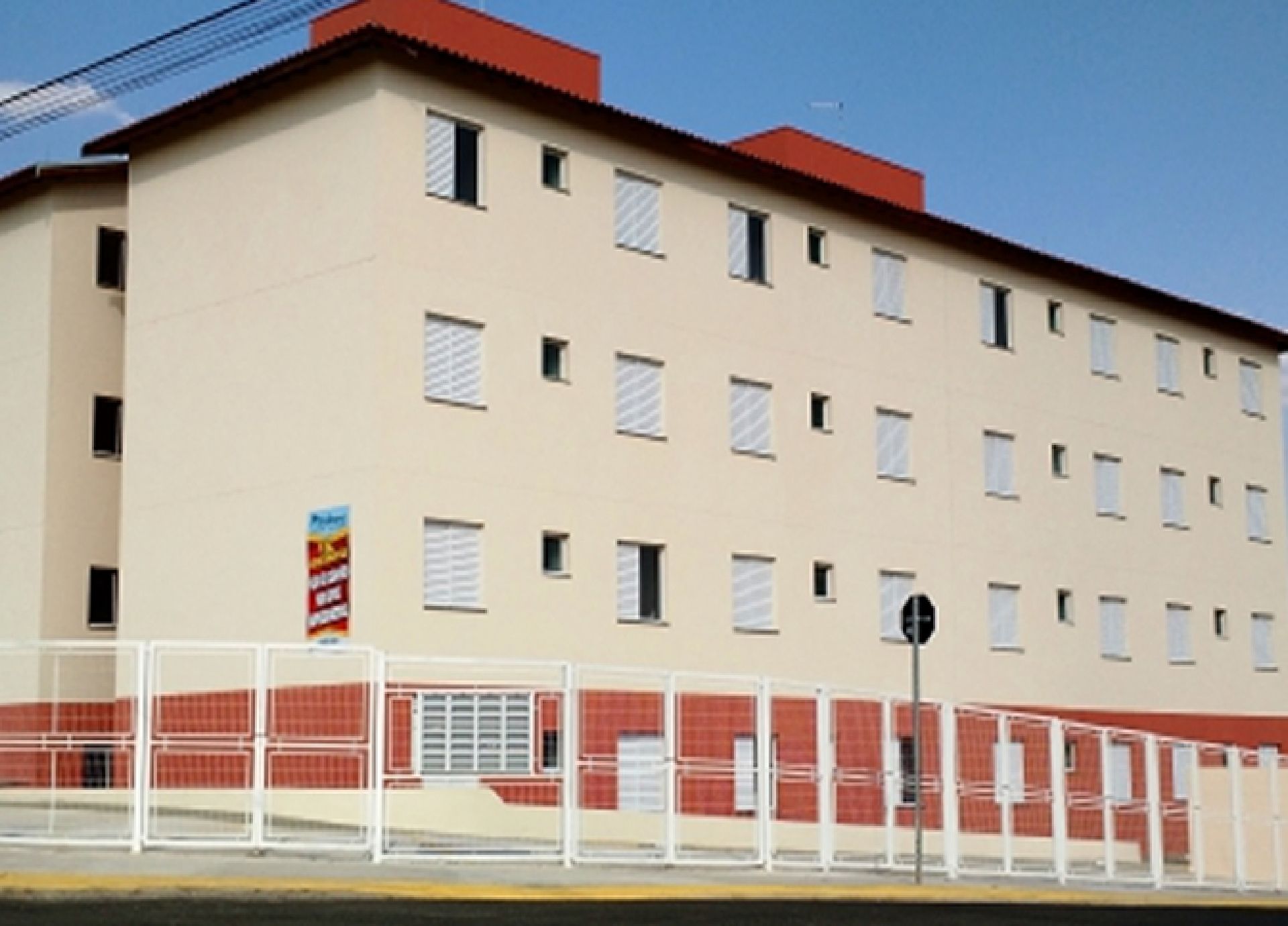 Prédio de apartamentos bege de vários andares com persianas brancas e telhado e cerca vermelhos.