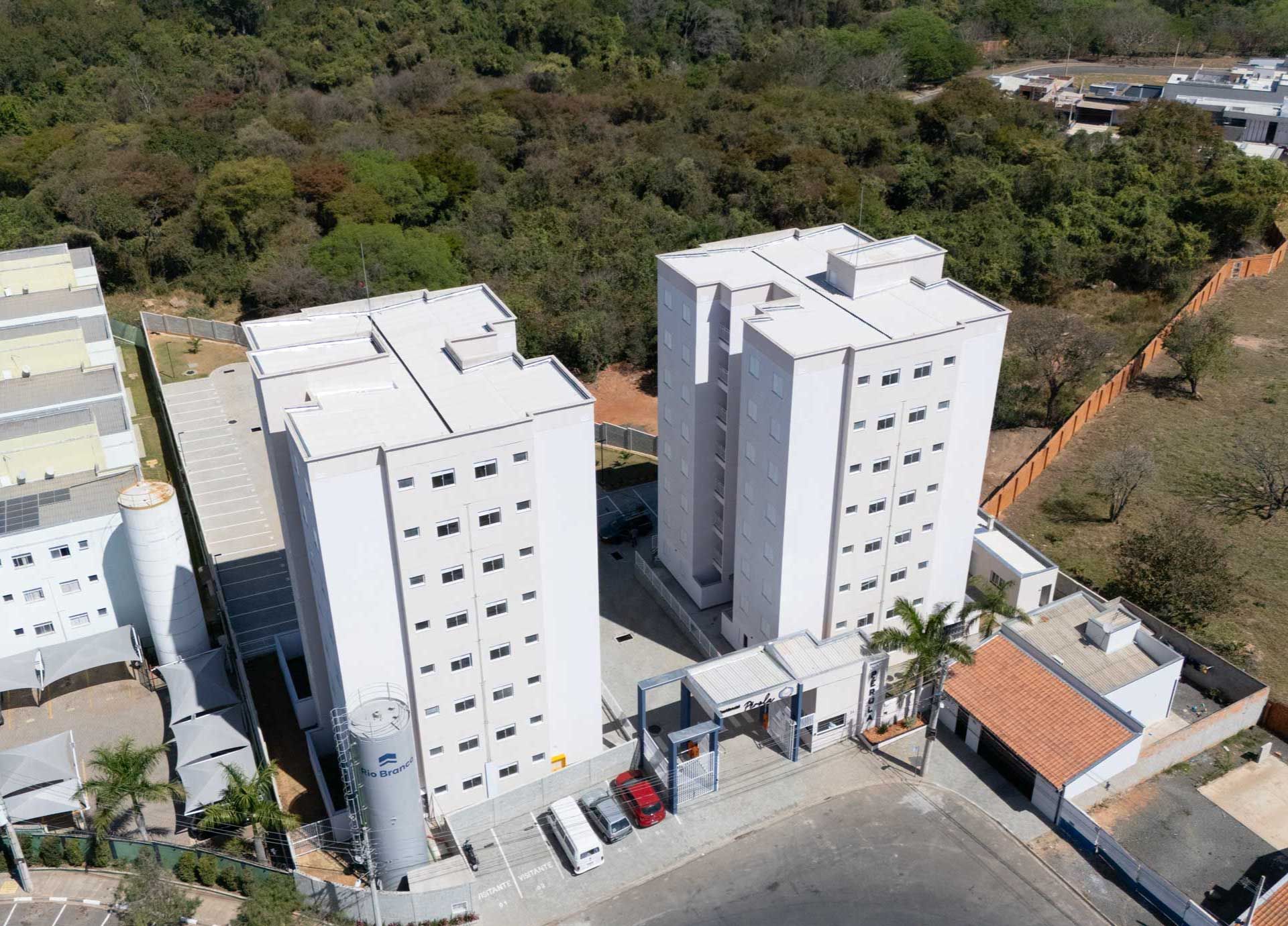 Vista aérea de muitos prédios altos e cinzas de apartamentos em um complexo, com piscina, quadra esportiva e área verde.