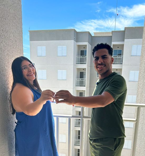 Duas pessoas em uma sacada sorrindo, brindando e olhando uma para a outra com um prédio ao fundo.