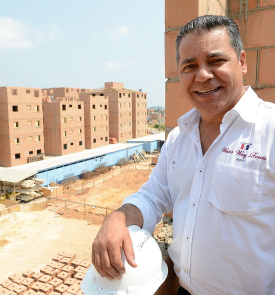 Homem de camisa branca, sorrindo, segurando um capacete, olhando para um prédio de apartamentos de tijolos em construção.