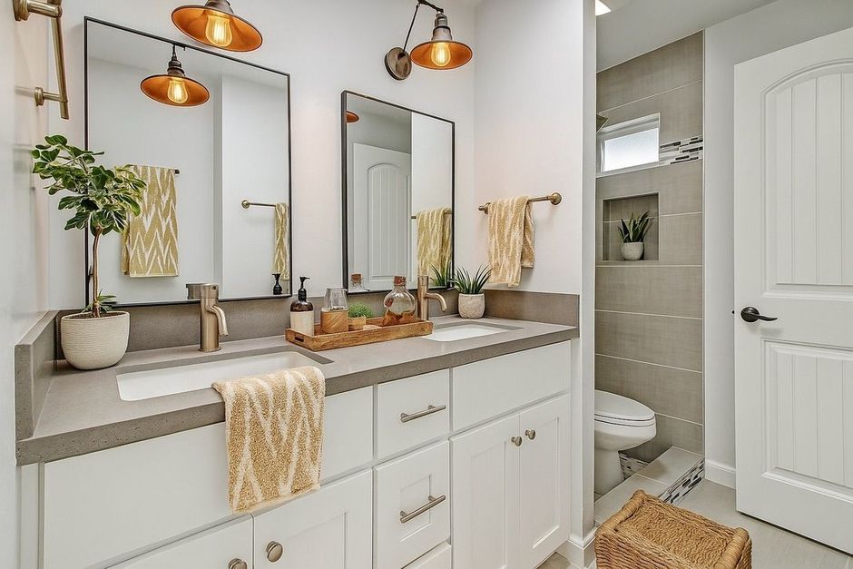 A bright bathroom with a double vanity, mirrors, and a toilet; light gray and white with gold accents.