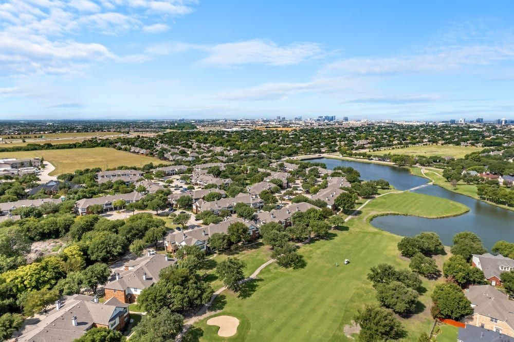 Aerial view of a suburban neighborhood with numerous houses closely packed together, adjacent to a golf course with green fairways and a large pond. In the background, a city skyline can be observed under a blue sky scattered with a few clouds at Marquis at Stonebriar in Frisco, TX.