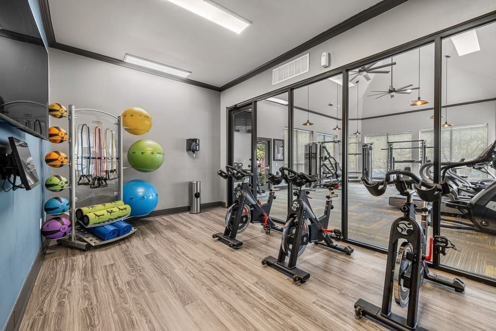 The interior of a well-equipped gym featuring stationary bikes in the foreground, weight training machines in the back, exercise balls, and resistance bands on the left, all under bright lighting in a clean, modern space with mirrors and large windows at Marquis at Stonebriar in Frisco, TX.
