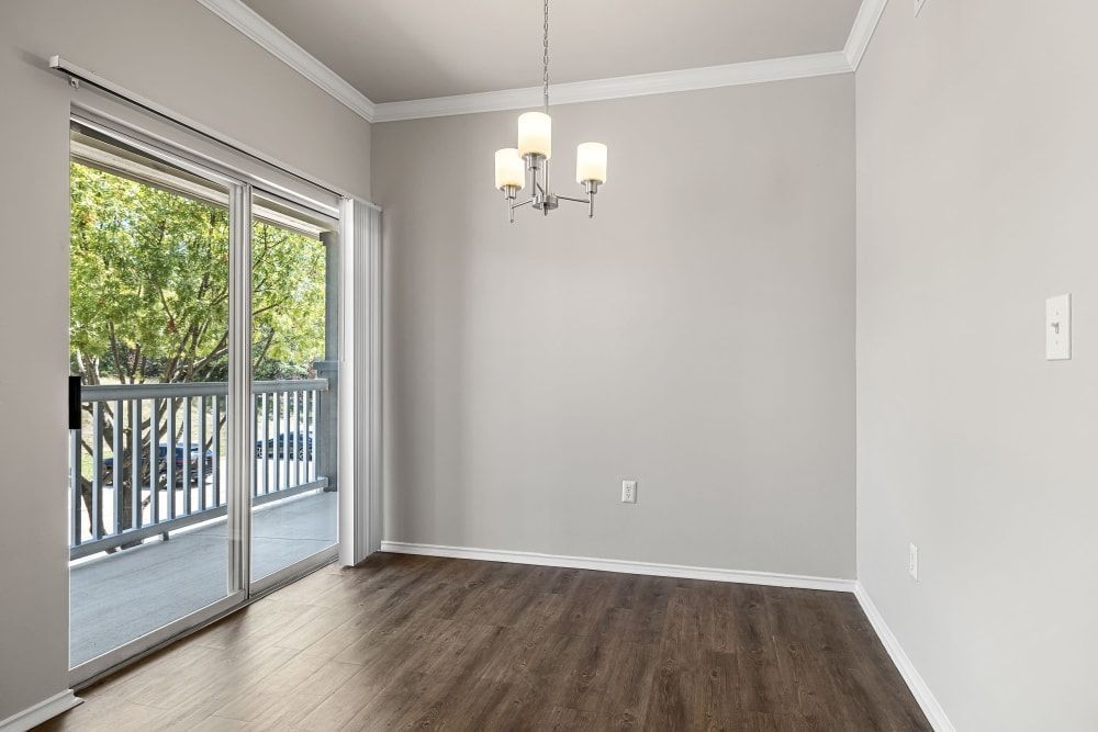 Empty residential room with light gray walls and dark wood flooring, featuring a sliding glass door leading to a balcony with a view of trees, and a simple white chandelier hanging from the ceiling at Marquis at Stonebriar in Frisco, TX.
