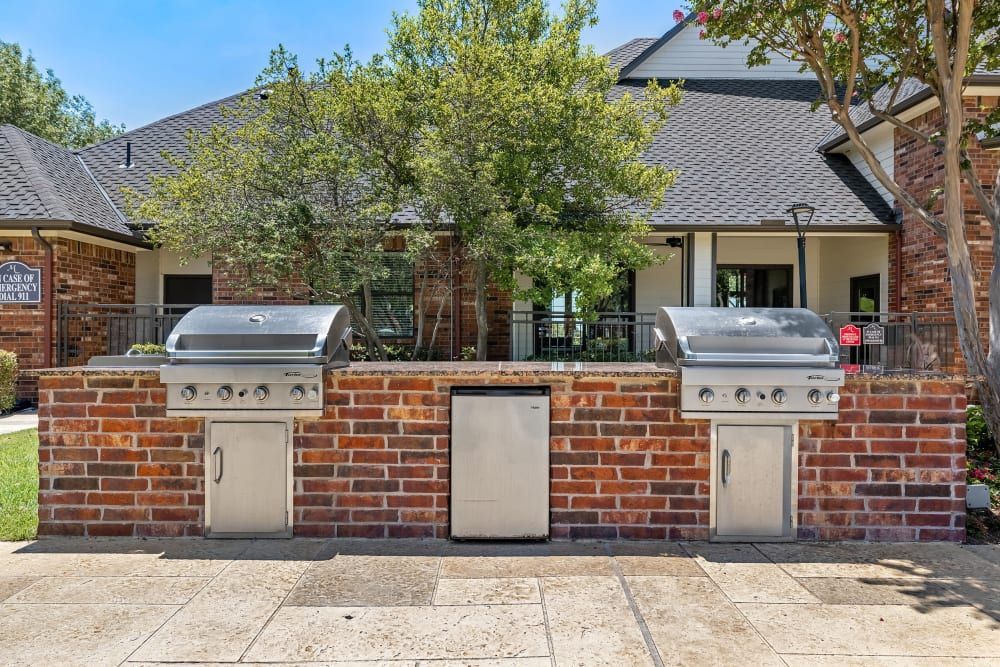 Outdoor brick grilling stations with two stainless steel gas grills built into a red brick counter, situated on a concrete patio in front of a suburban brick house with trees and a clear sky in the background at Marquis at Stonebriar in Frisco, TX.