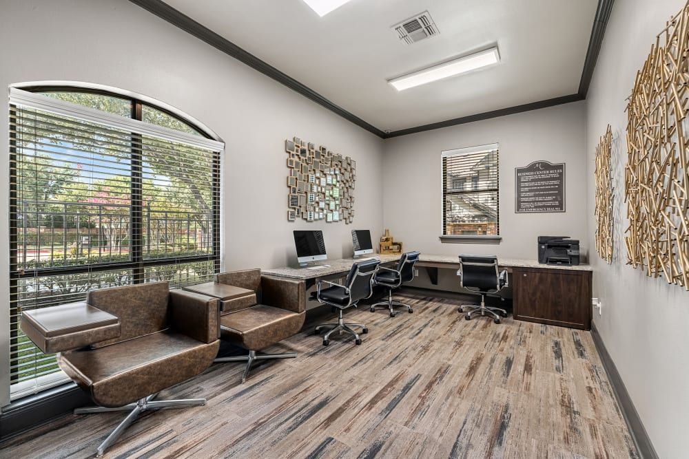 Modern and well-lit business center with a large window letting in natural light, stylish brown armchairs, a long desk with multiple computer workstations, and contemporary wall art decorations at Marquis at Stonebriar in Frisco, TX.