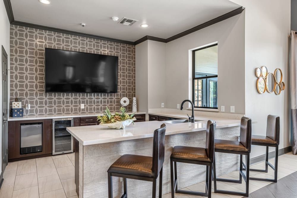 Modern apartment community kitchen interior with a geometric patterned backsplash, dark wood cabinets, and a white countertop island featuring black bar stools. A wall-mounted flat-screen TV is visible above the cabinets, and decorative mirrors hang beside a window on the adjacent wall at Marquis at Stonebriar in Frisco, TX.