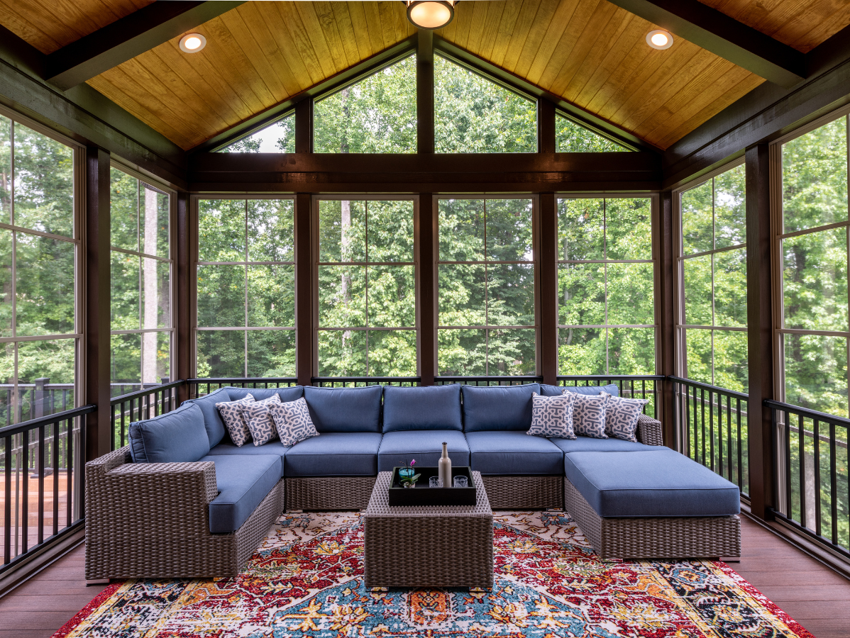 Screened porch with blue sectional, patterned rug, and a wooded view.
