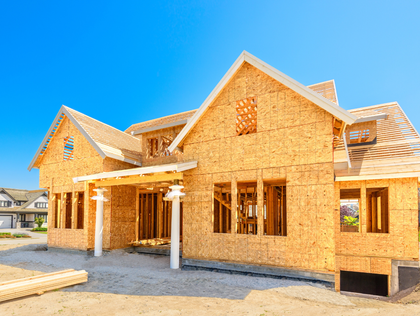 House under construction, wood frame visible against a bright blue sky.