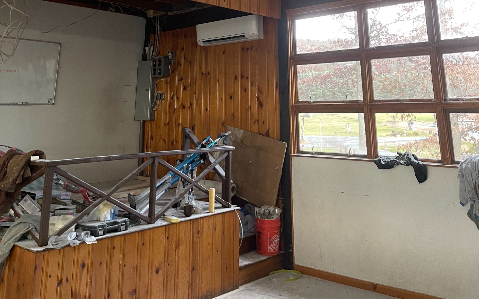 Interior room with wooden paneling, a small balcony, and a window overlooking a yard; cluttered with tools and materials.