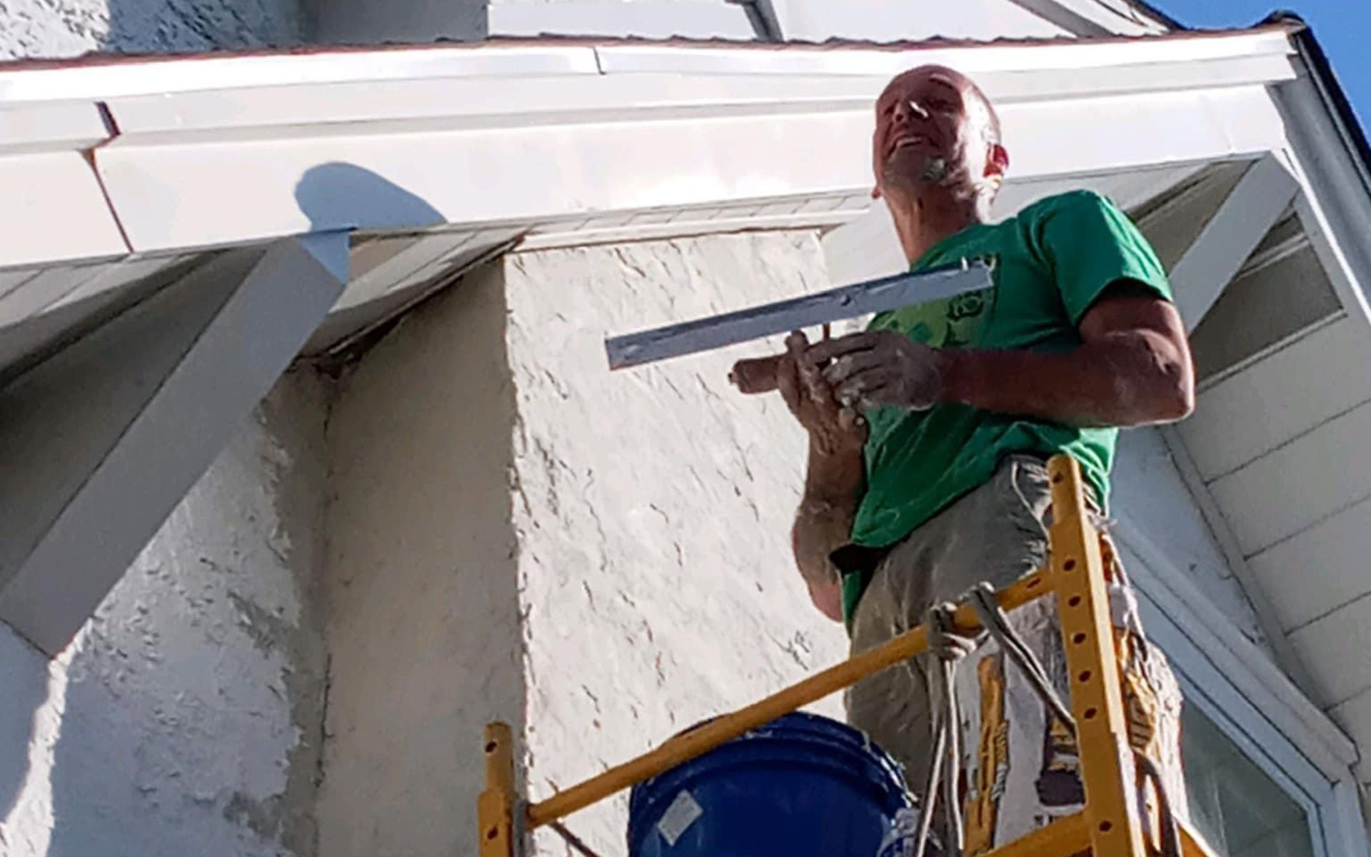 Man on scaffold applying stucco with a trowel to a chimney, next to white trim.