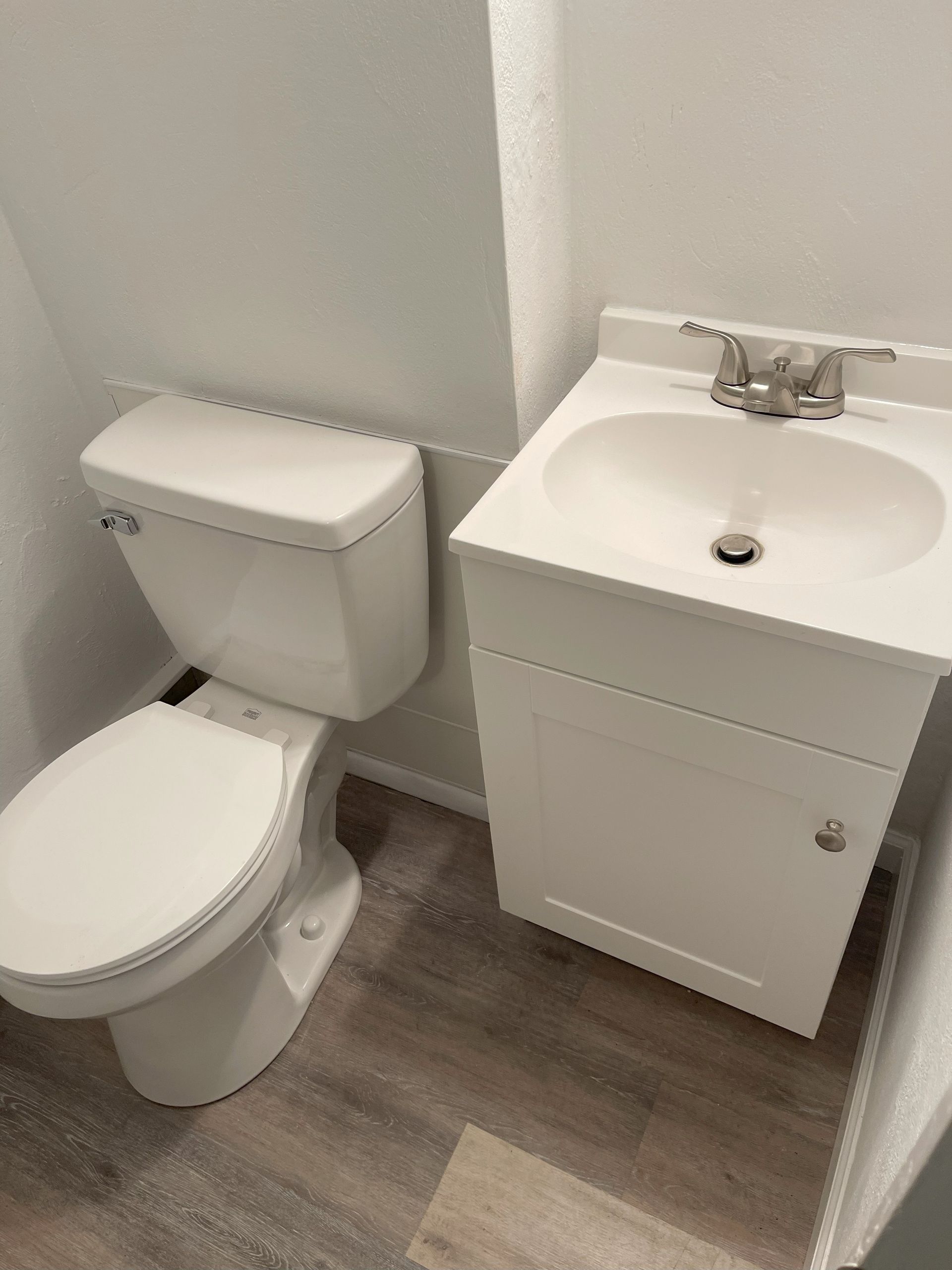 White toilet and sink in a small bathroom with wooden floors and white walls.