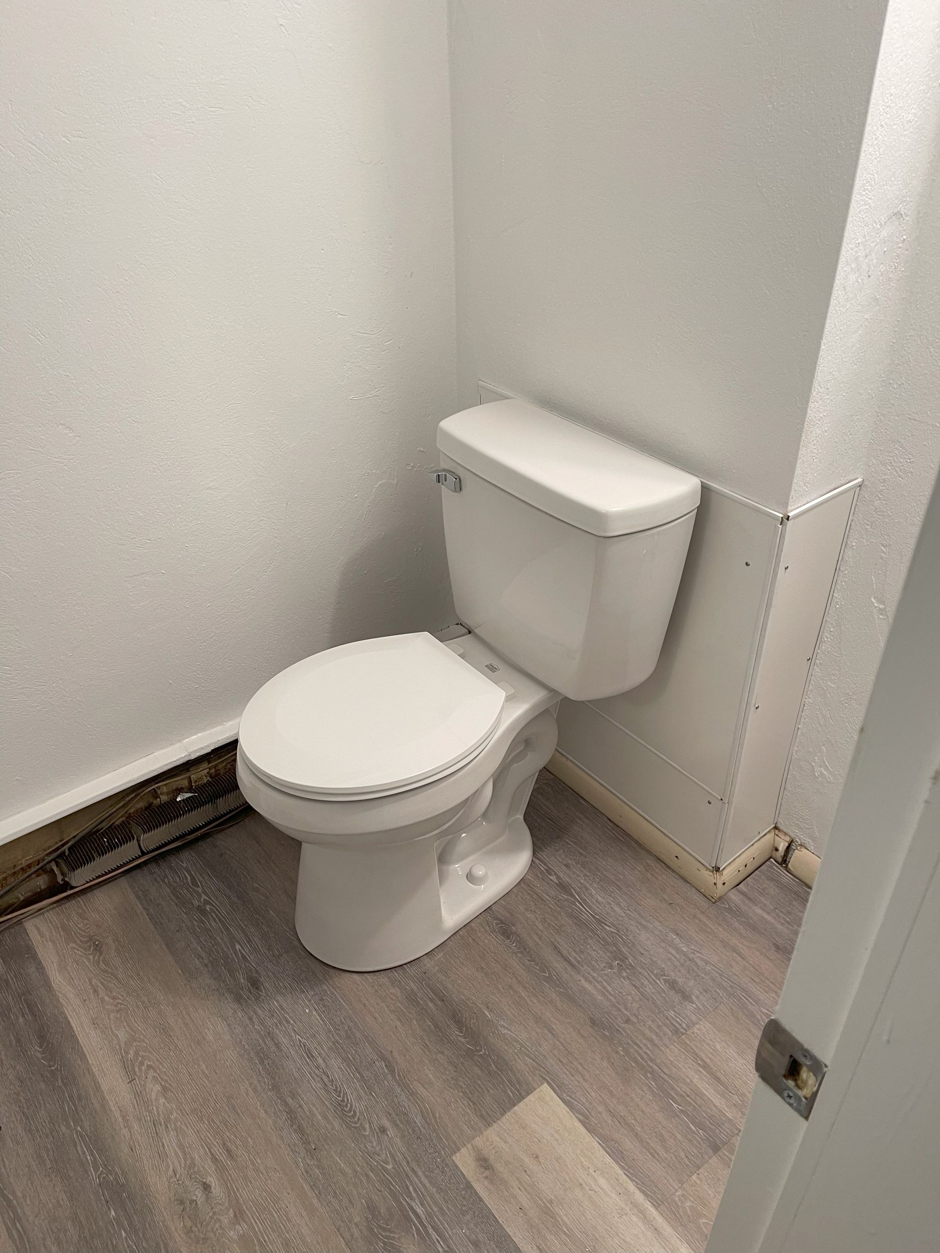 White toilet in a small bathroom with gray wood-look flooring and white walls.