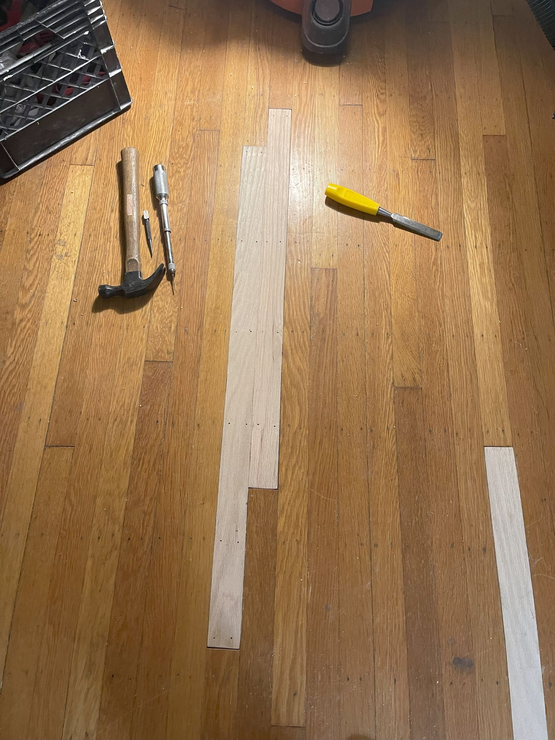Tools (hammer, screwdriver, chisel) on wooden floor with light-colored strips, likely for repair.